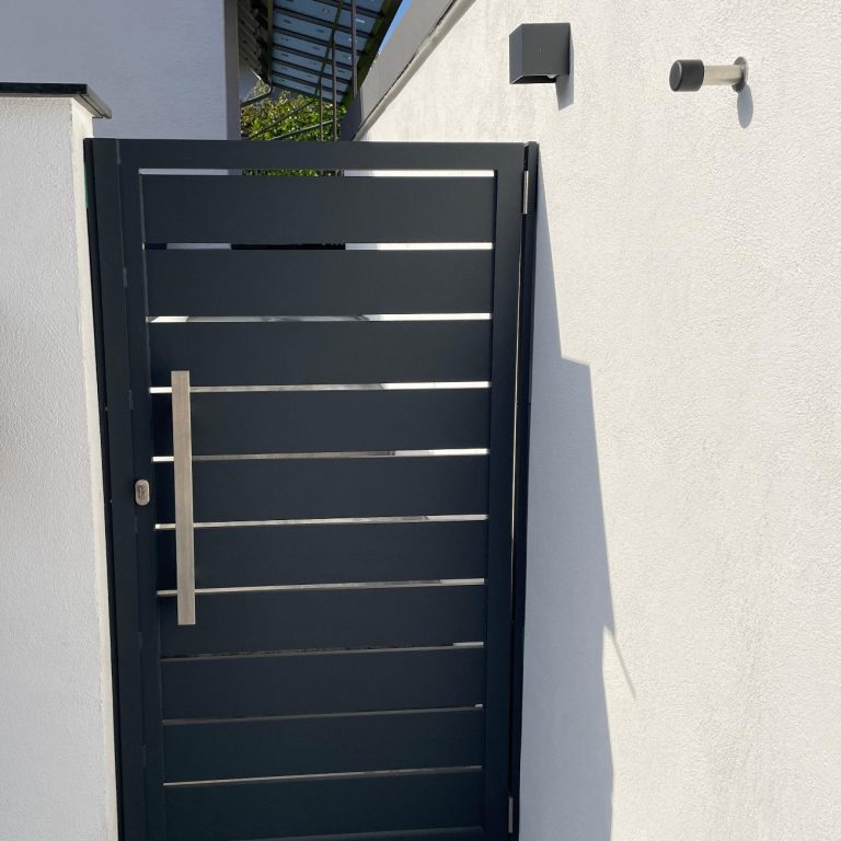 Gartentüre Aluminium verschweißt und Pulverbeschichtet mit E-Öffner Langlebig Individuell Ästethisch Edelstahl Beschläge Schwarze Aluminiumtür mit horizontaler Füllung und moderner Griffstange sowie Türstopper an einer weißen Wand.