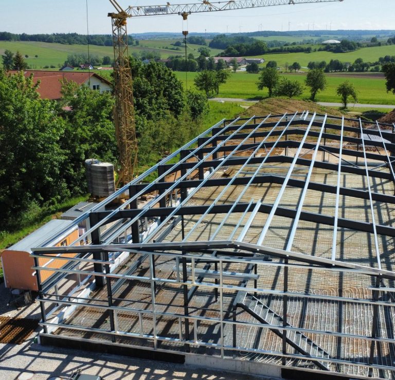 Hallenkonstruktion Stahl, Büroeinbau mit Treppe und Geländer Pfetten Wandriegel Stahlbau Hallenbau Blick auf ein Baustellengelände mit Stahlhalle , Treppe, Technikbühne und grüner Landschaft im Hintergrund.